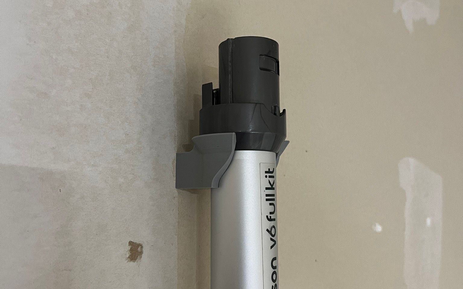 A grey printed bracket screwed to an unfinished plaster wall, cradling the aluminium tube of a cordless vacuum below its dark grey collar.