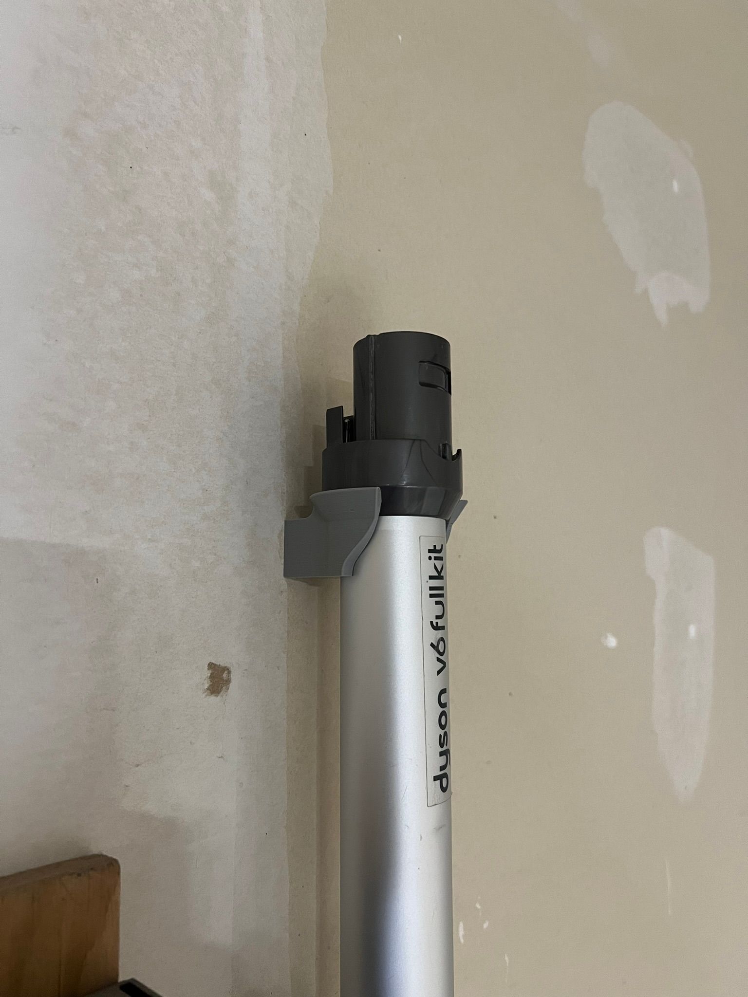 A photograph of a grey printed bracket screwed to an unfinished plaster wall, cradling the aluminium tube of a cordless vacuum just below its dark grey collar. A printed label on the tube reads dyson v6 full kit.
